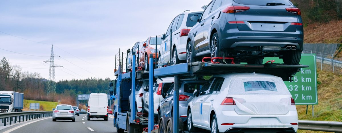 A car carrier truck, part of an Open Car Transport Dallas service, transports multiple new SUVs on a highway, surrounded by other vehicles. Road signs and trees are visible along the roadside under a cloudy sky.