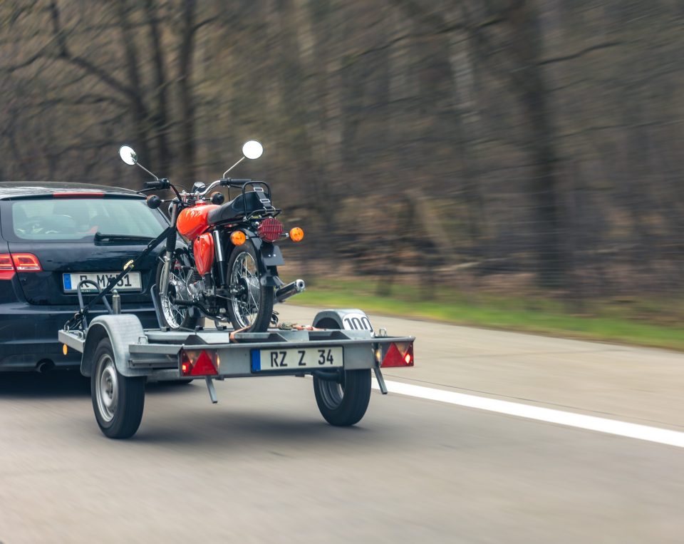 A black car tows a trailer carrying a red motorcycle on a highway, with blurred trees in the background suggesting movement—a scene reminiscent of Open Car Transport Dallas services, expertly navigating the roads for reliable vehicle shipping.