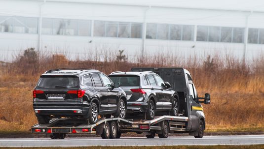 A black truck transports two black SUVs on a car trailer along a road, with dry grass and an industrial building in the background—a common scene for Auto Transport Dallas TX services.