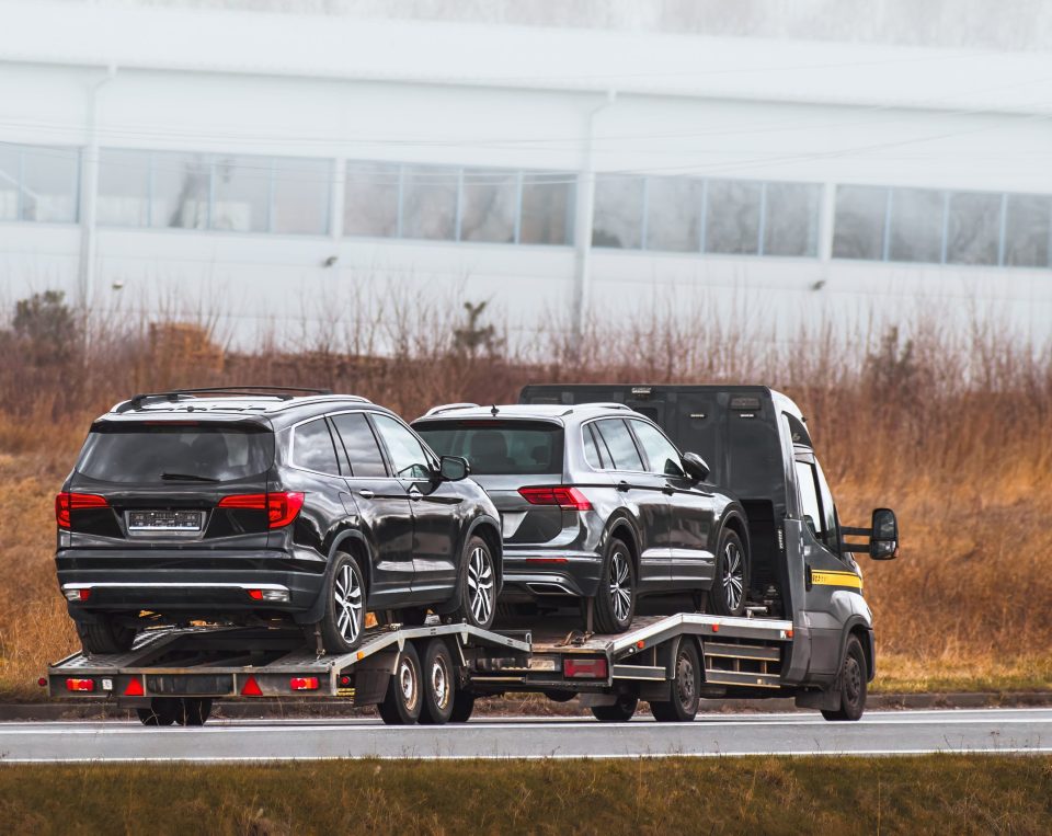 A black truck transports two black SUVs on a car trailer along a road, with dry grass and an industrial building in the background—a common scene for Auto Transport Dallas TX services.